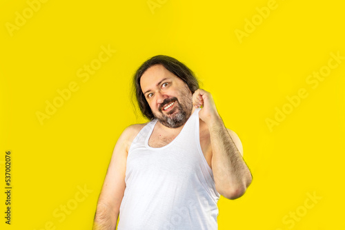 A contented unshaven man in a white undershirt holding his shirt hysterically smiling. Unemployed slovenly dressed man from Eastern Europe. © Николай Батаев