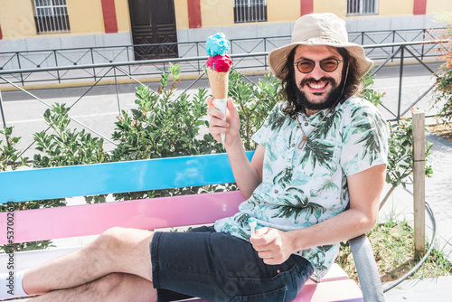 Person eating ice cream. A young bearded hipster rests lying on a bench on a hot day in Seville. Man enjoying the taste of a two-flavor waffle cone. Male in vintage style with sunglasses and hat