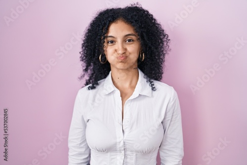 Hispanic woman with curly hair standing over pink background puffing cheeks with funny face. mouth inflated with air, crazy expression.