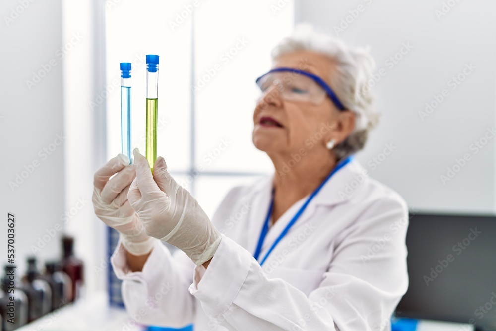 Senior grey-haired woman wearing scientist uniform holding test tubes at laboratory Senior grey-haired woman wearing scientist uniform holding test tubes at laboratory
