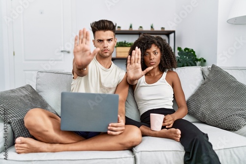 Young interracial couple using laptop at home sitting on the sofa doing stop sing with palm of the hand. warning expression with negative and serious gesture on the face.