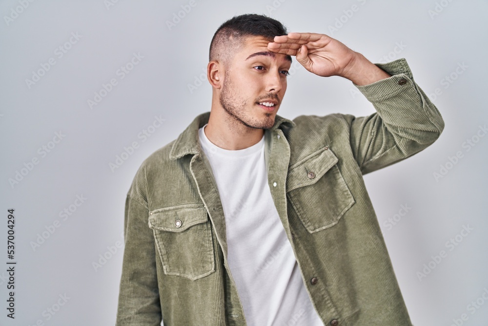 Young hispanic man standing over isolated background very happy and smiling looking far away with hand over head. searching concept. Young hispanic man standing over isolated background very happy and smiling looking far away with hand over head. searching concept.
