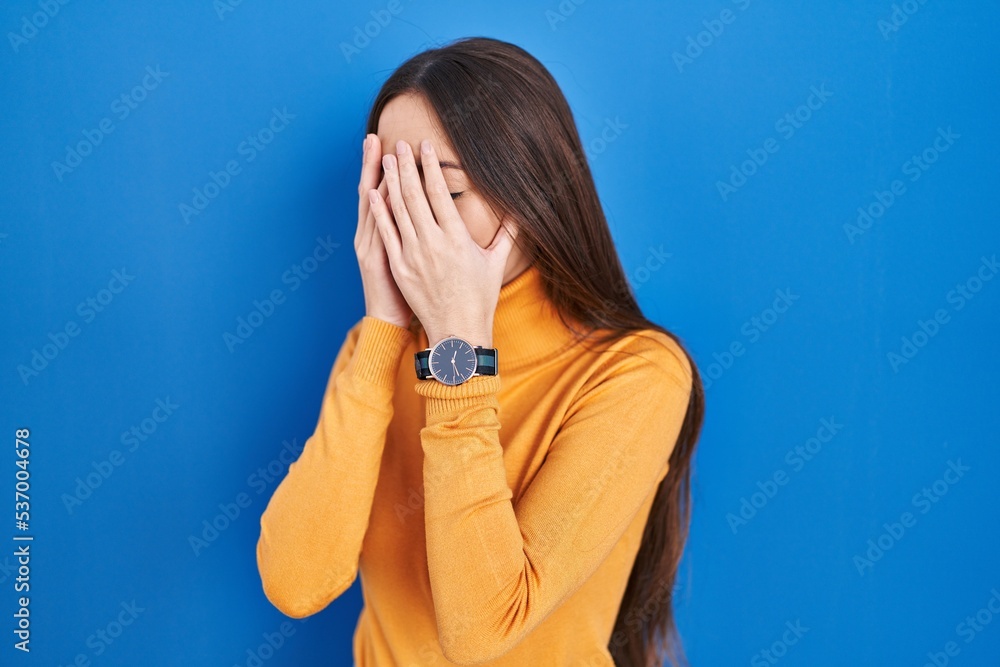 Young brunette woman standing over blue background with sad expression covering face with hands while crying. depression concept. Young brunette woman standing over blue background with sad expression covering face with hands while crying. depression concept.
