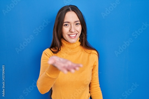 Young brunette woman standing over blue background smiling cheerful offering palm hand giving assistance and acceptance.