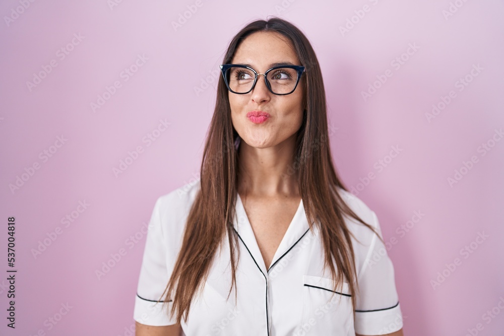 Young brunette woman wearing glasses standing over pink background smiling looking to the side and staring away thinking.
