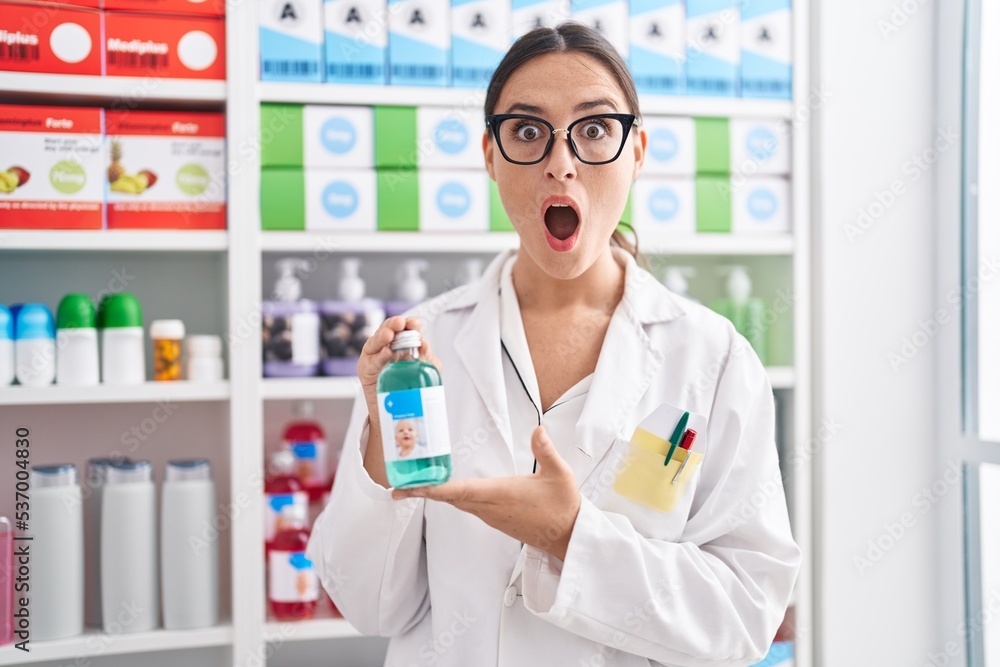Brunette woman working at pharmacy drugstore holding cough syrup afraid and shocked with surprise and amazed expression, fear and excited face.