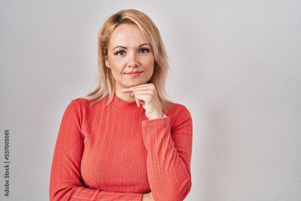 Blonde woman standing over isolated background with hand on chin thinking about question, pensive expression. smiling and thoughtful face. doubt concept. Blonde woman standing over isolated background with hand on chin thinking about question, pensive expression. smiling and thoughtful face. doubt concept.