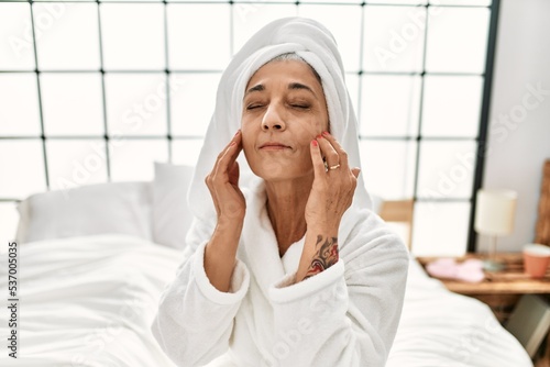 Middle age grey-haired woman massaging face with cream sitting on bed at bedroom