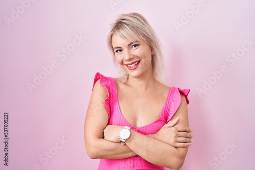 Young caucasian woman standing over pink background happy face smiling with crossed arms looking at the camera. positive person.