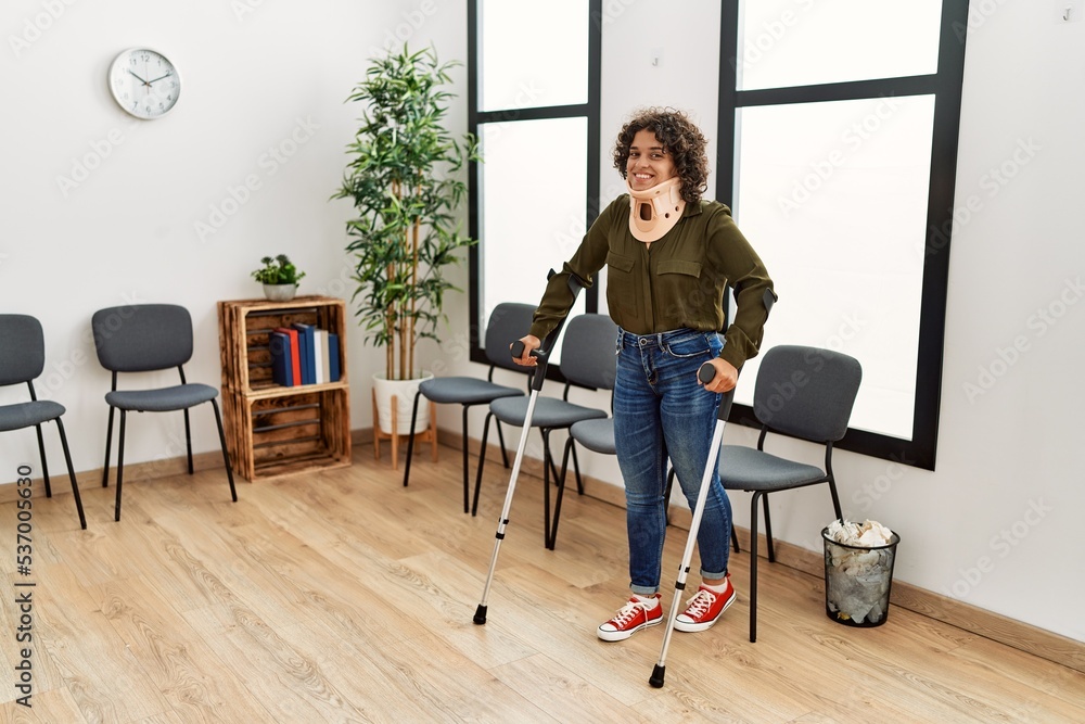 Young hispanic woman wearing cervical collar walking using crutches at clinic waiting room