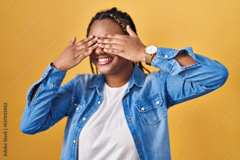 African american woman with braids standing over yellow background covering eyes with hands smiling cheerful and funny. blind concept. African american woman with braids standing over yellow background covering eyes with hands smiling cheerful and funny. blind concept.