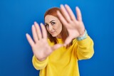 Young woman standing over blue background doing frame using hands palms and fingers, camera perspective