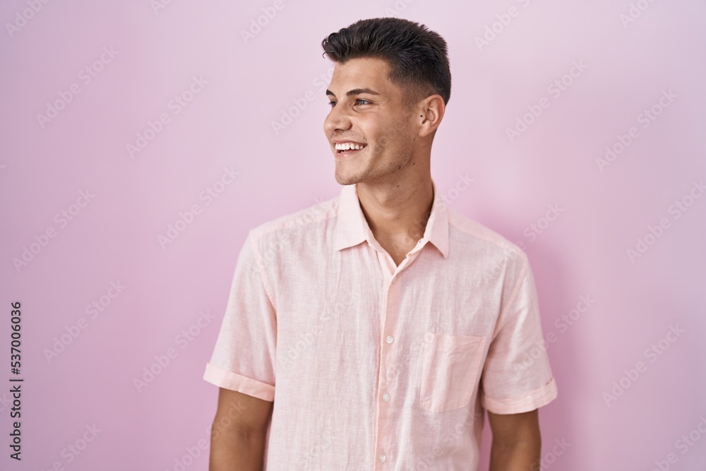 Young hispanic man standing over pink background looking away to side with smile on face, natural expression. laughing confident. Young hispanic man standing over pink background looking away to side with smile on face, natural expression. laughing confident.