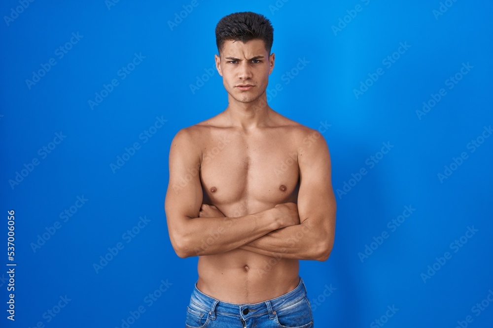 Young hispanic man standing shirtless over blue background skeptic and nervous, disapproving expression on face with crossed arms. negative person.