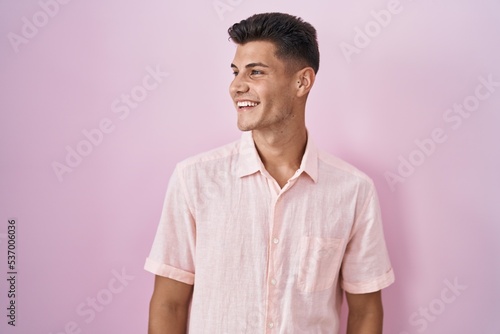 Young hispanic man standing over pink background looking away to side with smile on face, natural expression. laughing confident.