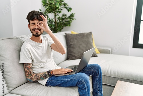 Hispanic man with beard sitting on the sofa doing ok gesture with hand smiling, eye looking through fingers with happy face.