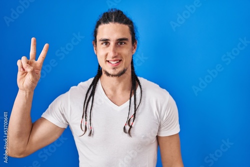 Hispanic man with long hair standing over blue background showing and pointing up with fingers number two while smiling confident and happy.