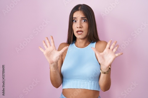 Young brunette woman standing over pink background afraid and terrified with fear expression stop gesture with hands, shouting in shock. panic concept. © Krakenimages.com
