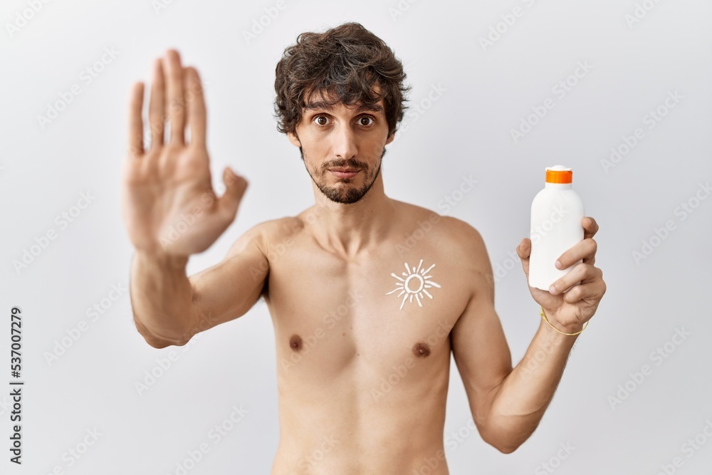 Young hispanic man standing shirtless holding sunscreen lotion doing stop sing with palm of the hand. warning expression with negative and serious gesture on the face.