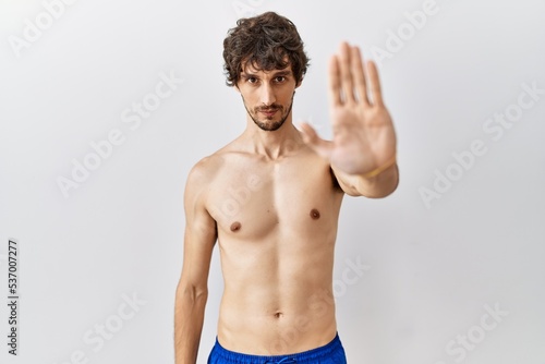 Young hispanic man standing shirtless over isolated, background doing stop sing with palm of the hand. warning expression with negative and serious gesture on the face.