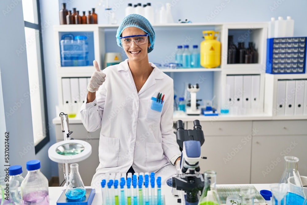 Brunette woman working at scientist laboratory doing happy thumbs up gesture with hand. approving expression looking at the camera showing success. Brunette woman working at scientist laboratory doing happy thumbs up gesture with hand. approving expression looking at the camera showing success.