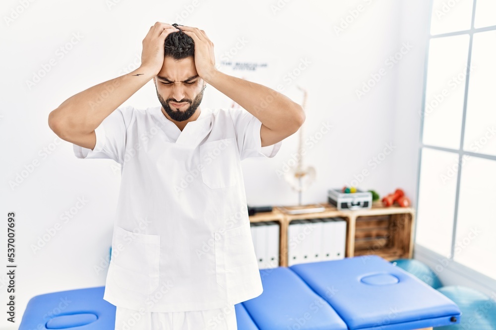 Obraz premium Young handsome man with beard working at pain recovery clinic suffering from headache desperate and stressed because pain and migraine. hands on head.
