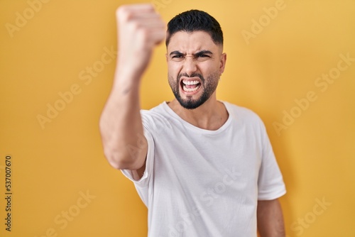 Young handsome man wearing casual t shirt over yellow background angry and mad raising fist frustrated and furious while shouting with anger. rage and aggressive concept.