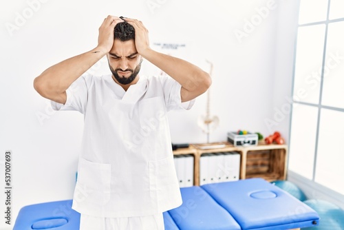 Young handsome man with beard working at pain recovery clinic suffering from headache desperate and stressed because pain and migraine. hands on head.
