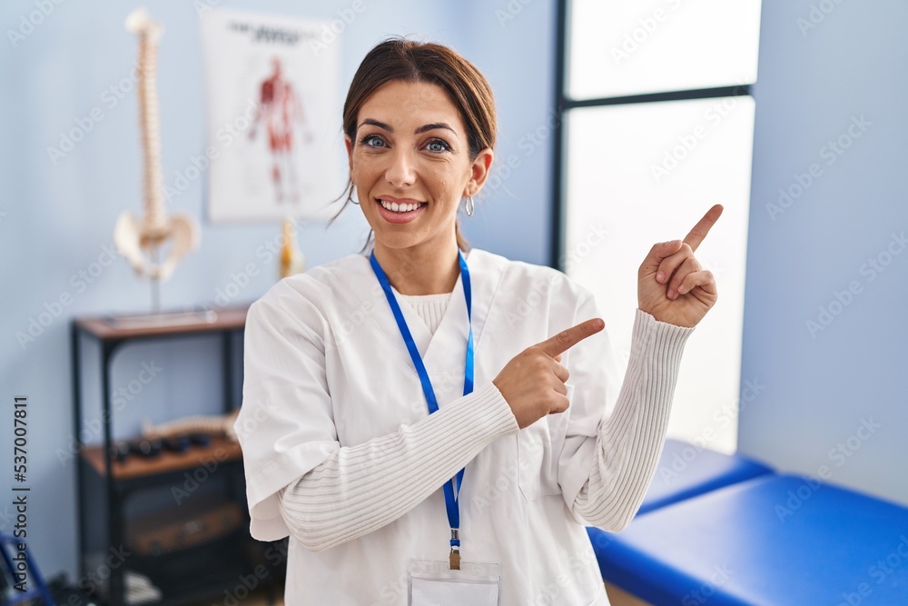 Young brunette woman working at pain recovery clinic smiling and looking at the camera pointing with two hands and fingers to the side. Young brunette woman working at pain recovery clinic smiling and looking at the camera pointing with two hands and fingers to the side.