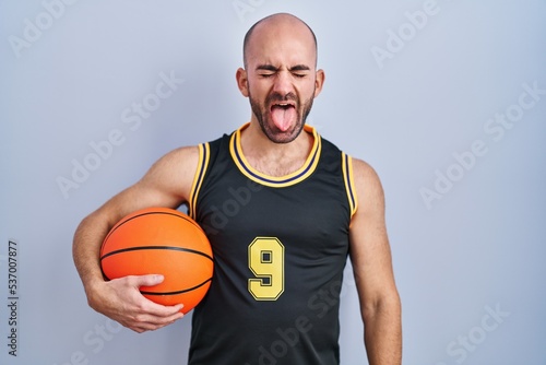 Young bald man with beard wearing basketball uniform holding ball sticking tongue out happy with funny expression. emotion concept.