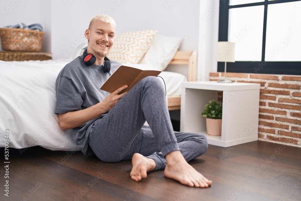 Young caucasian man reading book sitting on floor at bedroom