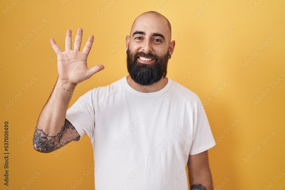Young hispanic man with beard and tattoos standing over yellow background showing and pointing up with fingers number five while smiling confident and happy. Young hispanic man with beard and tattoos standing over yellow background showing and pointing up with fingers number five while smiling confident and happy.