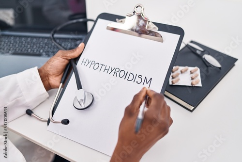 African american woman wearing doctor uniform writing hypothyroidism on clipboard at clinic photo