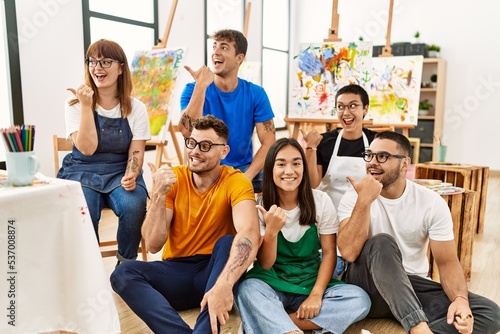 Group of people sitting at art studio pointing thumb up to the side smiling happy with open mouth