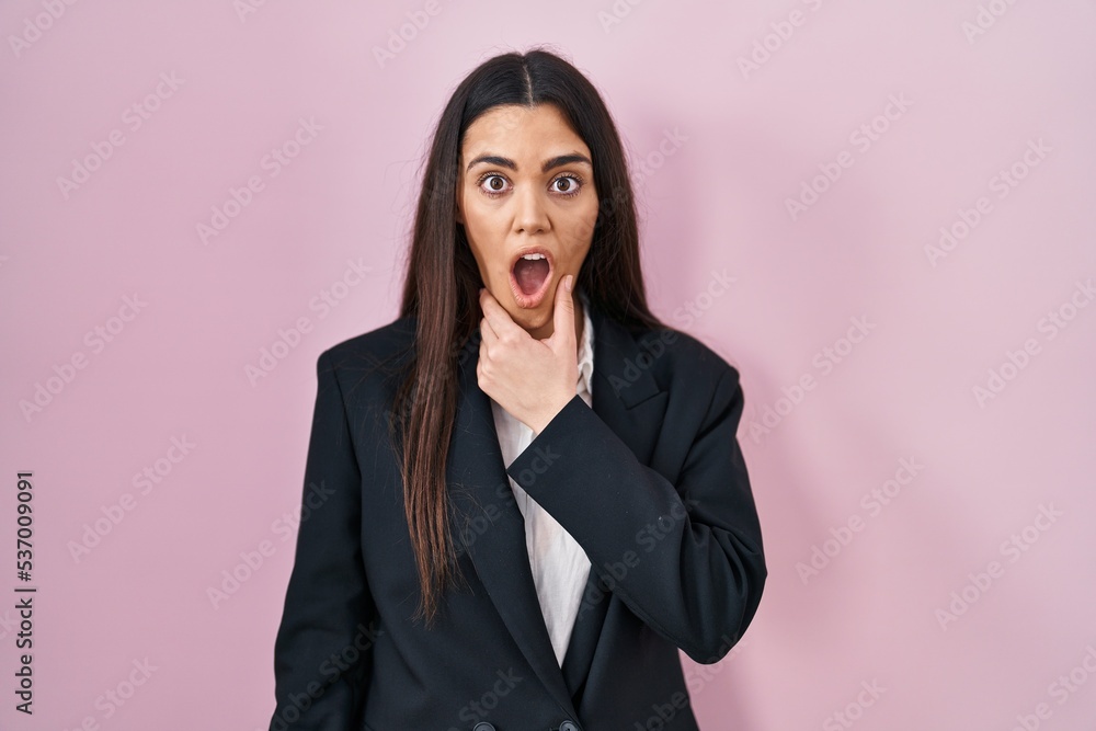 Young brunette woman wearing business style over pink background looking fascinated with disbelief, surprise and amazed expression with hands on chin Young brunette woman wearing business style over pink background looking fascinated with disbelief, surprise and amazed expression with hands on chin