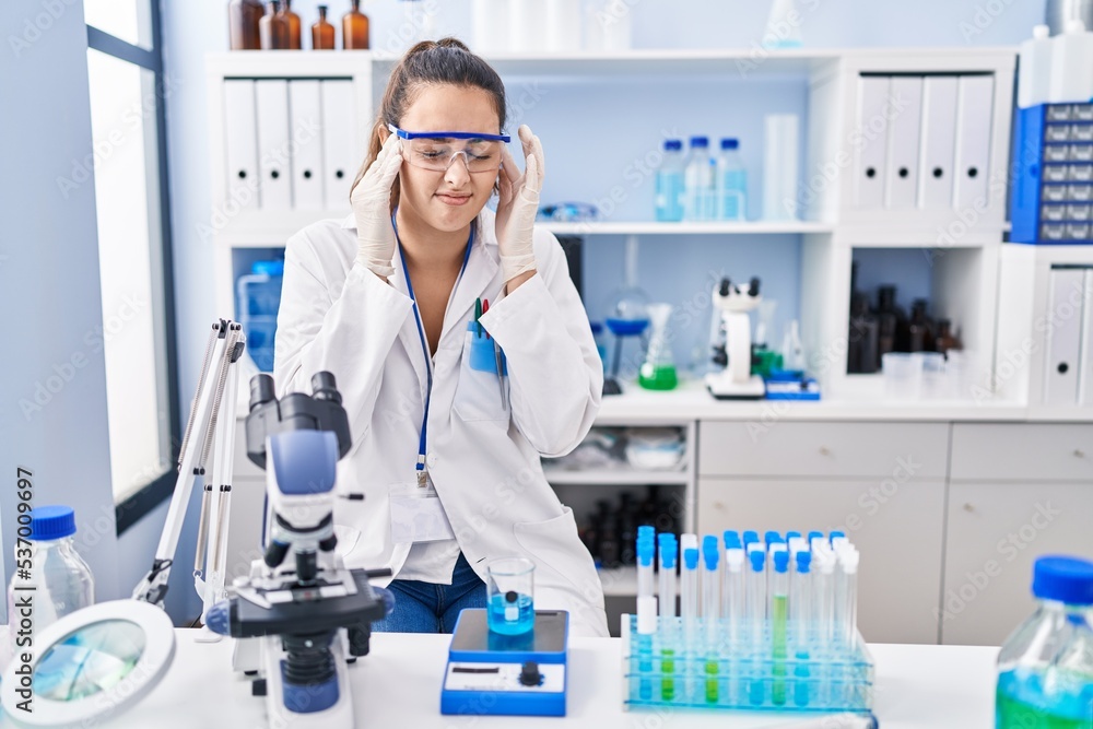 Young hispanic woman working at scientist laboratory with hand on head for pain in head because stress. suffering migraine. Young hispanic woman working at scientist laboratory with hand on head for pain in head because stress. suffering migraine.