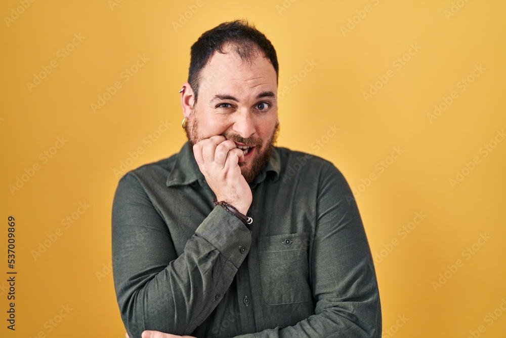 Plus size hispanic man with beard standing over yellow background looking stressed and nervous with hands on mouth biting nails. anxiety problem.