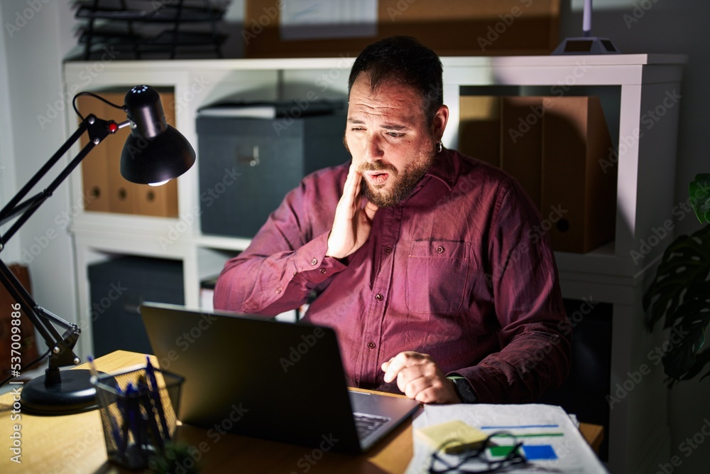 Plus size hispanic man with beard working at the office at night touching mouth with hand with painful expression because of toothache or dental illness on teeth. dentist Plus size hispanic man with beard working at the office at night touching mouth with hand with painful expression because of toothache or dental illness on teeth. dentist