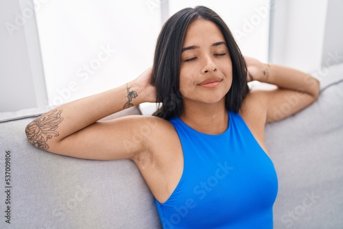 Young hispanic woman relaxed with hands on head sitting on sofa at home