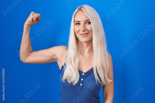 Caucasian woman standing over blue background strong person showing arm muscle, confident and proud of power