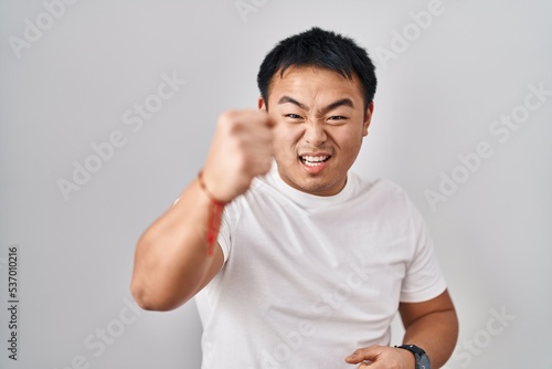 Young chinese man standing over white background angry and mad raising fist frustrated and furious while shouting with anger. rage and aggressive concept.