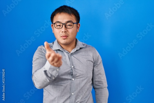 Young chinese man standing over blue background looking at the camera blowing a kiss with hand on air being lovely and sexy. love expression.
