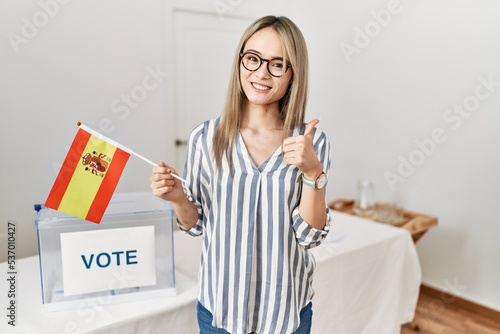 Asian young woman at political campaign election holding spain flag smiling happy and positive, thumb up doing excellent and approval sign