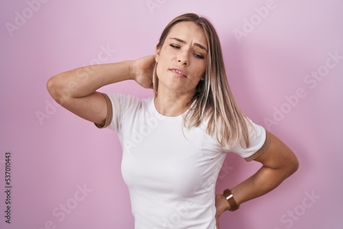 Blonde caucasian woman standing over pink background suffering of neck ache injury, touching neck with hand, muscular pain