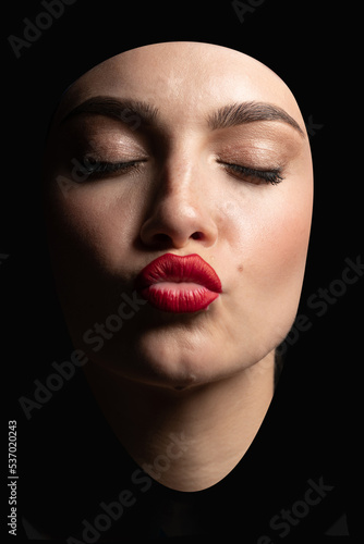 Beauty, fashion and make-up concept. Abstract portrait of a young and beautiful woman with closed eyes sending kiss isolated over black background. Model with sun tan, classic make-up and red lipstick