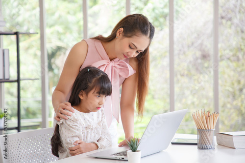 Children education and home school concept : Young asian mother pleased to see little daughters' study online. Serious child girl while online learning with pleasant mom at home. © aekkorn