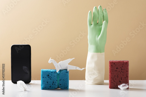 Composition with smartphone sponges and rubber glove on beige background. Improve common housework with cleaning service. Easy solution for chores (ID: 537022022)