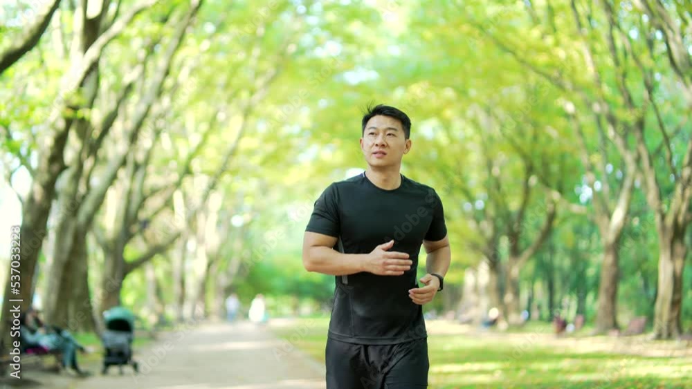 sporty young asian runner running on alley in city urban park. Morning jogging of an athletic man outdoors in nature. Sport Male athlete jogger in black sportswear, slow motion sporty young asian runner running on alley in city urban park. Morning jogging of an athletic man outdoors in nature. Sport Male athlete jogger in black sportswear, slow motion