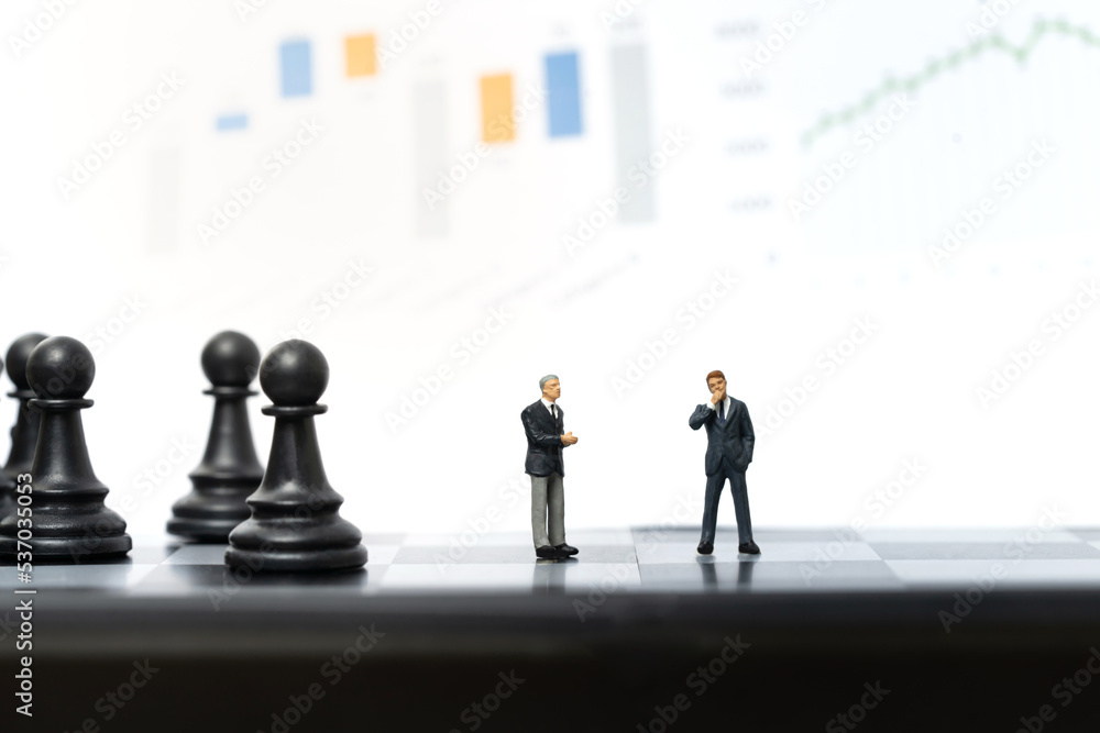 Miniature people, two businessmen stand on chessboard with graph on background. Business and financial concept Miniature people, two businessmen stand on chessboard with graph on background. Business and financial concept