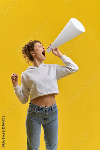 Side view of pretty girl standing up with megaphone. Young female with curly blonde hair shouting, crying, raising fist with closed eyes. Concept of freedom and human rights.
